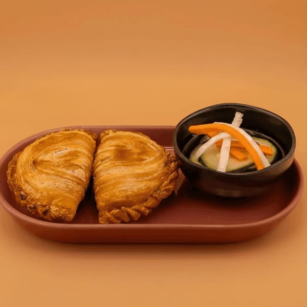 Two golden-brown curry puffs and a small bowl of pickled vegetables on a reddish-brown plate.