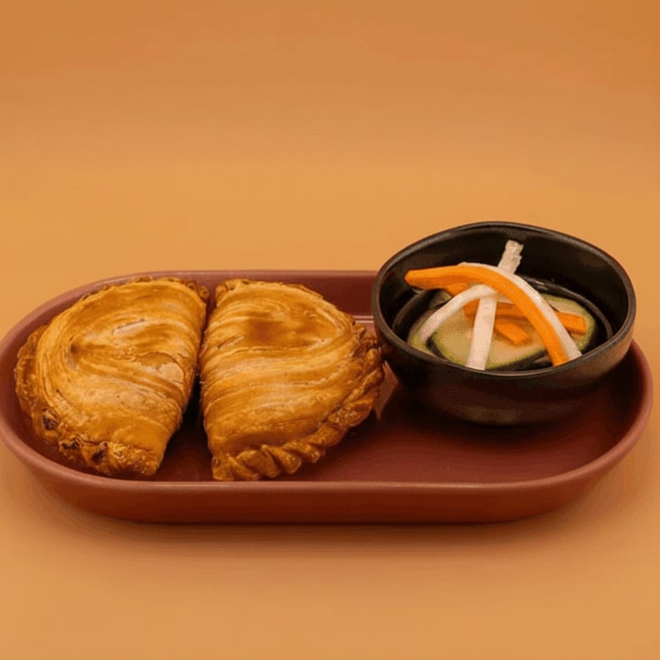 Two golden-brown curry puffs and a small bowl of pickled vegetables on a reddish-brown plate.