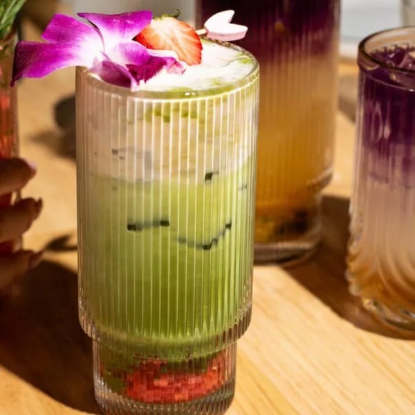 Green iced drink with foam, a strawberry slice, and a purple orchid in a ribbed glass.