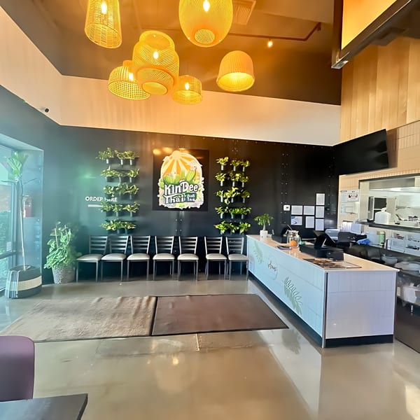 The interior of Kin Dee Thai restaurant features a counter, waiting chairs, and hanging plants.