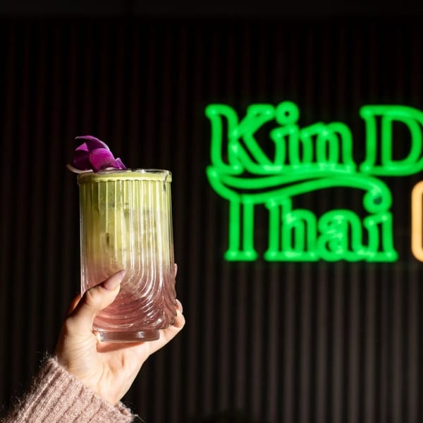 Hand holding a green cocktail garnished with a purple orchid, with a neon Kin Dee Thai sign in the background.