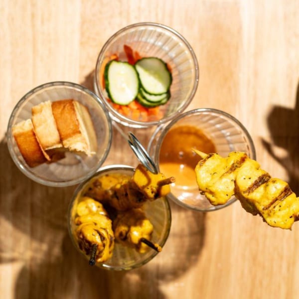 Kin Dee Thai chicken satay skewers, peanut sauce, pickled cucumbers, and toast on a wooden table.